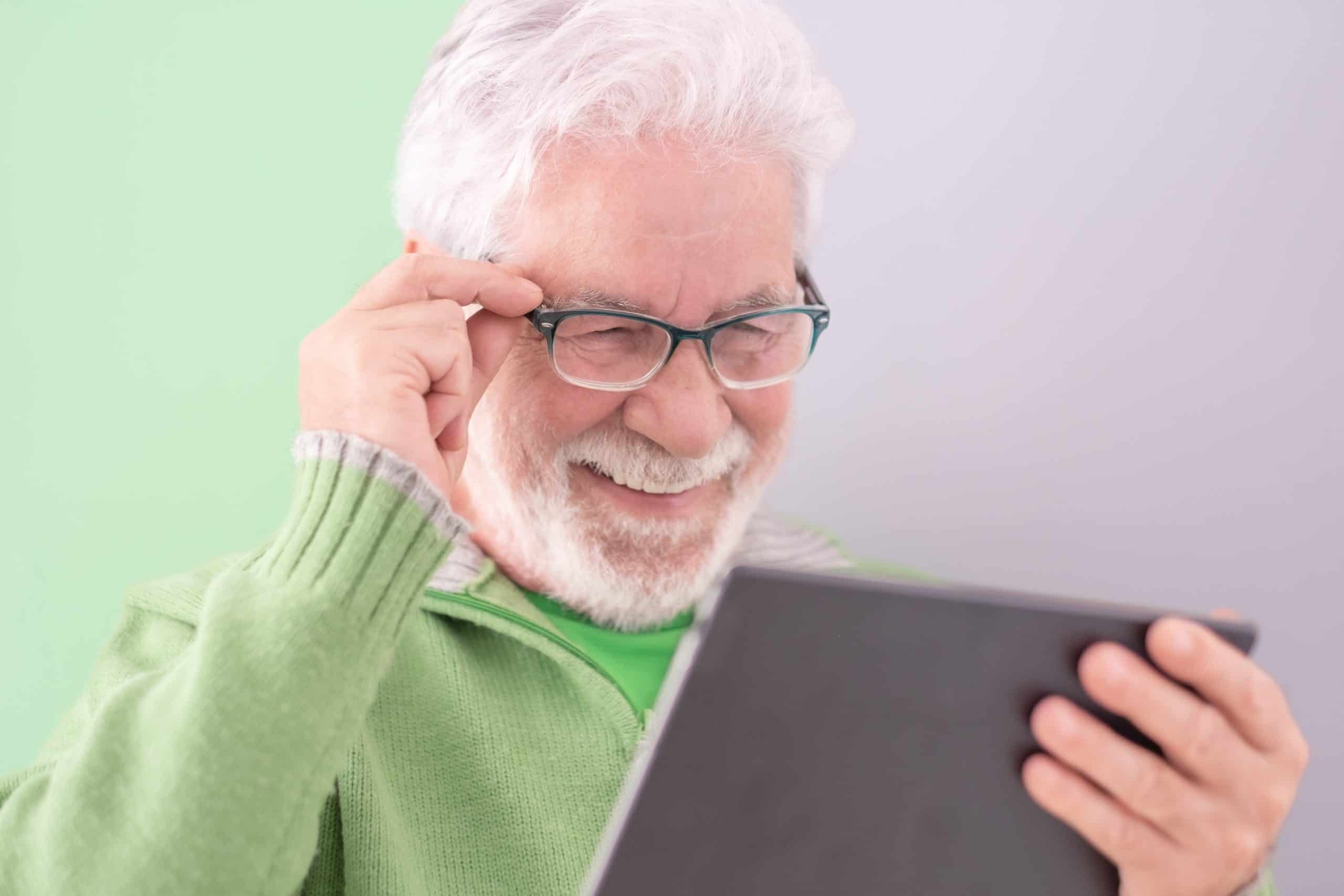 Smiling confident male teacher with a tablet-pc held in his hands standing on cute background. White-haired mature caucasian man with glasses, college student of the third age university